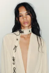 Camisa color crema o marfil, de tejido semitransparente, con escote pico cruzado y mangas largas con puños abotonados. Presenta un detalle de foulard incorporado en el frente, adornado con apliques de abalorios oscuros en forma de motivos florales y geométricos.