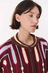 Campera tipo bomber tejida con diseño jacquard geométrico en tonos azul marino, blanco, rojo y verde. Presenta cierre frontal completo, cuello, puños y ruedo acanalados en color verde y blanco, con corte oversized y hombros caídos.