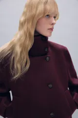 Abrigo corto de paño con mezcla de lana, color bordó. Presenta cuello subido, cierre frontal cruzado con botones, mangas largas con trabillas decorativas y bolsillos laterales de vivo.