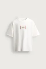 Camiseta boxy fit de color blanco con cuello redondo y mangas cortas. Presenta un parche rectangular en el centro del pecho con texto impreso y costuras decorativas a contraste en los laterales.