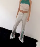 Pantalón de cuero sintético metalizado plateado, de corte recto con terminación acampanada y aberturas en los tobillos. Presenta una intervención artística con pintura abstracta en tonos rosados y verdes en la parte inferior.