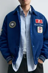 Cazadora estilo varsity confeccionada en pana de algodón azul, con interior ligeramente acolchado. Presenta cuello solapa, cierre frontal con botones a presión, bolsillos laterales de vivo y acabados en rib a rayas en puños y cintura. Incluye múltiples parches bordados decorativos en el frente.