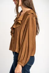Blusa de color marrón con acabado satinado, cuello en V con volados y lazo de ajuste frontal. Presenta mangas largas con puños elásticos y un corte holgado.