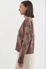 Cardigan de punto con escote en V y cierre frontal de botones. Presenta un diseño de estampado argyle en tonos marrones con líneas blancas. Posee mangas largas con puños acanalados y ruedo también acanalado.