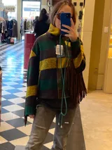 Campera de lana italiana con diseño de rayas horizontales en tonos verde, amarillo y marrón. Presenta flecos largos de gamuza en las mangas, cierre frontal y bolsillos laterales. La prenda se encuentra completamente forrada.