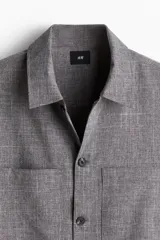 Sobrecamisa gris de tejido texturizado, con cuello inglés, tapeta lisa con botones oscuros y dos bolsillos superiores abiertos. Presenta un corte recto y holgado.