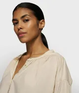Blusa oversize de algodón orgánico color beige claro, con cuello redondo y cierre parcial de botones. Presenta detalles fruncidos en hombros, pecho y espalda, y puños de punto jersey.