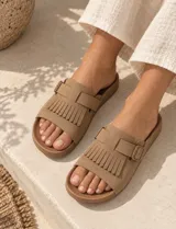 Sandalias de gamuza color marrón, con diseño de una sola tira ancha decorada con flecos y una hebilla metálica lateral ajustable. Cuenta con plantilla anatómica.