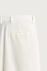Pantalón relaxed fit de color blanco off-white, confeccionado en tejido con lino. Cuenta con pliegues frontales en la cintura, bolsillos delanteros y cierre frontal con cremallera y botón.
