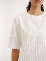 Blusa blanca de manga corta y cuello redondo, con diseño de rayas verticales texturizadas y detalles sutiles de flecos.