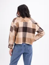 Sobrecamisa de corte cropped con diseño a cuadros en tonos rosa y beige. Presenta cuello clásico, cierre frontal con botones metálicos, mangas largas con puños abotonados y dos bolsillos frontales con solapa.