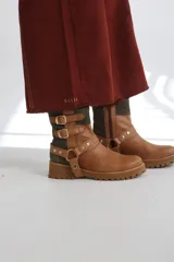 Bota estilo biker de caña media, confeccionada en cuero negro con detalles en gamuza marrón. Presenta correas ajustables con hebillas metálicas, tachas decorativas y suela track dentada.