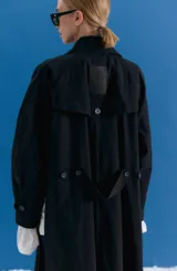 Trench midi de color negro, confeccionado en una mezcla de algodón y nylon con propiedades repelentes al agua. Presenta un diseño holgado con tajos laterales, bolsillos internos y detalle de etiqueta en la espalda.
