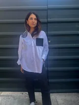 Camisa blanca de corte clásico con bordado de hilo negro en el hombro y bolsillo de cuerina negro en el pecho.