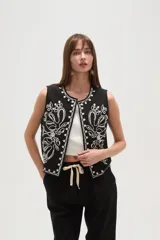 Chaleco negro sin mangas, de corte corto y abierto, con bordado floral en hilo blanco que recorre el borde del cuello y los frentes.