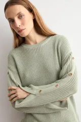 Sweater de punto color blanco natural, con cuello redondo y mangas largas adornadas con botones dorados a lo largo de las costuras exteriores.