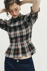 Blusa de cuello subido con diseño de cuadros en tonos marrones, negros y blancos. Presenta mangas cortas con terminación en volantes y un corte ajustado con detalle de volado en la cintura.