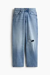 Jean de corte wide leg con diseño de cinco bolsillos, confeccionado en denim de algodón rígido. Presenta un lavado claro con efecto desgastado y un detalle de rotura en la rodilla izquierda. Cuenta con cintura clásica, cierre de botón y bragueta.