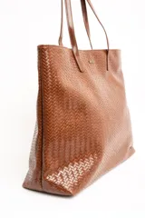 Bolso tipo shopper color marrón de cuero sintético con diseño trenzado. Cuenta con doble asa de hombro y placa metálica con el logo de la marca en el frente.
