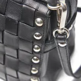 Cartera tipo baúl pequeña de cuero negro, con cuerpo tejido en patrón de cuadros (intrecciato) y tachas metálicas redondas en los bordes. Posee doble asa superior y correa ajustable para llevar al hombro o cruzada.