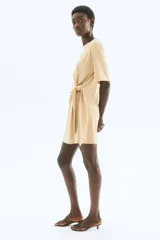 Vestido corto de punto color beige, con cuello redondo y mangas cortas. Presenta un nudo fruncido en el lateral que crea un efecto drapeado y abertura en el ruedo.