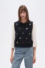Chaleco de punto color azul marino, con cuello V y diseño de rombos trenzados. Presenta múltiples apliques bordados de motivos variados como mariposas, cerezas, flores, soles, lazos, limones, fresas, sonrisas, rosas y trozos de pizza.