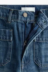 Pantalón de denim azul con lavado medio, corte de pierna recta y tiro alto. Presenta bolsillos de parche delanteros con solapa, trabillas para cinto y detalles de pliegues marcados en el centro de las piernas.