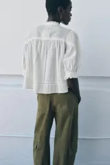 Camisa blanca de algodón con cuello redondo y manga por debajo del codo abullonada. Presenta detalles de bordados a tono y cierre frontal con botones.