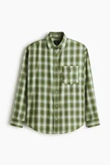 Camisa de manga larga de corte relajado, confeccionada en mezcla de algodón suave con estampado a cuadros en tonos verdes y blancos. Presenta cuello inglés, cierre frontal de botones, un bolsillo aplicado en el pecho y dobladillo redondeado.