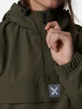 Canguro tipo anorak de color gris oscuro, con capucha ajustable, bolsillo frontal con solapa y cierre de media cremallera. Presenta detalles en color naranja en los tiradores de los cierres y un parche con el logo de la marca en el centro del pecho.