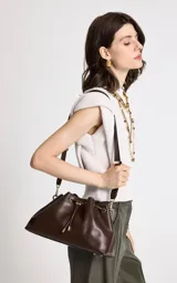 Cartera tipo cubo de Le Lis Blanc, color chocolate, con cierre superior mediante cordón ajustable y herrajes metálicos dorados. Cuenta con una correa larga ajustable para llevar al hombro o cruzada.