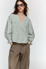 Blusa de escote pico y manga larga con puño elástico y volante. Presenta un diseño de rayas verticales finas en azul claro y blanco, con flores en relieve.