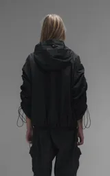 Campera rompevientos negra de calce oversize, confeccionada en microfibra técnica ligera con acabado semi-mate. Presenta capucha integrada, cierre frontal técnico y bolsillos tipo ojal. Destaca por su sistema de frunces elásticos regulables mediante tancas a lo largo de las mangas y el ruedo, permitiendo ajustar el volumen de la prenda.