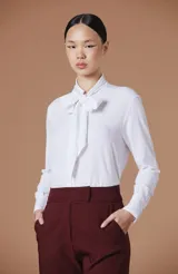 Blusa blanca de manga larga con cuello alto y detalle de lazo frontal tipo moño. Confeccionada en tejido ligero y fluido con corte entallado.