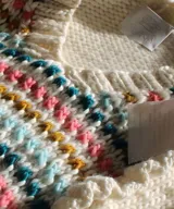 Chaleco de punto blanco color marfil con escote en V, tejido a mano en lana merino. Presenta detalles de pequeños vuelos en las sisas y franjas horizontales con bordados texturizados de colores vibrantes en el frente.