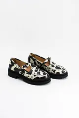 Mocasines estilo Mary Jane con diseño de estampado de vaca (cow print), confeccionados en cuero. Presentan una tira con hebilla ajustable sobre el empeine y una suela track negra de 4,5 cm de altura.
