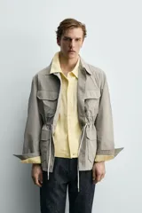 Parka relaxed fit color gris, confeccionada en tejido técnico ligero, con cuello solapa, manga larga con puño abotonado, cuatro bolsillos de plastrón con solapa en el frente, cintura ajustable con cordón y cierre frontal de botones.