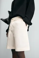 Bermuda de corte relajado color beige claro, confeccionada en tejido de algodón y lino. Presenta cintura con cordón ajustable del mismo tejido, bolsillos delanteros y bolsillos traseros de vivo. Cierre frontal con cremallera y botón.