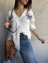 Camisa blanca de manga larga con escote en V y cierre frontal de botones. Presenta delicados bordados florales en tono oscuro en los hombros y puños.