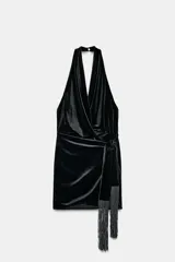 Vestido corto de terciopelo negro con cuello halter, escote en pico pronunciado y espalda descubierta. Presenta un detalle de nudo delantero con flecos largos y cierre de cremallera oculta.
