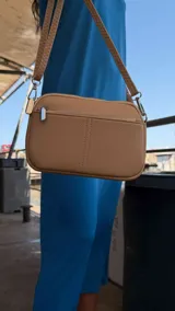 Bandolera pequeña de diseño rectangular en color beige, confeccionada en cuero sintético. Presenta una correa larga ajustable del mismo material y cierre superior con cremallera. Incluye el logo de la marca grabado en la parte frontal.