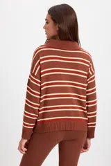 Campera de punto holgada con cuello tipo chomba y cierre frontal oculto. Presenta un diseño de rayas horizontales en tonos marrón, rojo y crema, con puños y ruedo acanalados. Incluye un parche bordado en la manga izquierda.