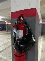 Bolso de hombro negro de charol, con forma alargada y cierre principal con cremallera. Presenta un dije metálico con forma de estrella de cuatro puntas en el frente y detalles de cremalleras decorativas en los laterales.