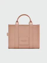 Cartera tipo tote de cuero granulado color tostado, con diseño estructurado y rectangular. Presenta doble asa superior fija y una correa larga ajustable y extraíble. En el frente, destaca el logo estampado con la inscripción 'THE TOTE BAG MARC JACOBS'.