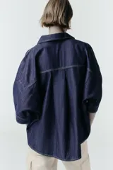 Camisa de denim azul oscuro con corte amplio, cuello solapa, escote en pico y mangas tres cuartos. Presenta un aplique metálico dorado en el centro y bajo asimétrico.