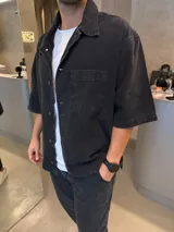 Camisa de jean de corte boxy y diseño holgado, color negro con efecto lavado. Presenta cuello clásico, cierre frontal con botones metálicos y un bolsillo frontal en el pecho.