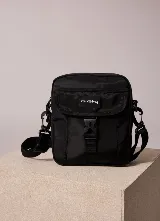 Bandolera Rusty modelo Vansi de color negro, con diseño estructurado, bolsillo frontal con solapa y cierre de hebilla plástica, correa regulable y removible, y etiqueta con el logo de la marca en el frente.