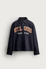 Sobrecamisa de ajuste relajado en color azul marino, con cuello de solapa, manga larga con puño abotonado y cierre frontal con botones. Presenta bordados contrastantes en el frente con la inscripción "St. Louis Worldwide Goods" en tonos naranja y blanco.