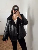 Campera tipo gamulán de cuero sintético negro con interior y solapas de corderito sintético al tono. Presenta cierre frontal, bolsillos laterales y detalles de hebillas en el cuello.