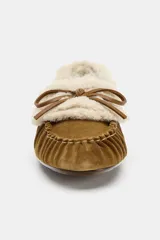 Mocasín plano tipo náutico confeccionado en serraje color tostado, con interior y borde superior forrados en tejido efecto borreguito. Presenta detalle fruncido en la puntera redonda y borlas decorativas en la parte frontal.